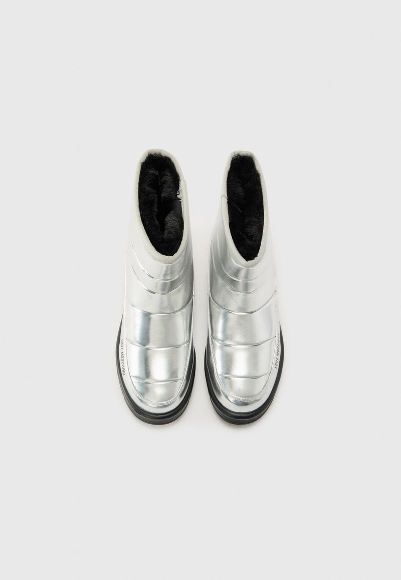 Bottines argentées avec un design brillant et rembourré, doublées de fausse fourrure noire. Elles disposent d'une semelle en caoutchouc noire et d'une fermeture éclair sur le côté.