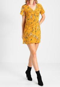 Keltainen kukkamekko, jossa lyhyet hihat, V-aukko ja istuva malli. Kuosissa valkoisia kukkia ja vihreitä lehtiä. Yhdistetty mustiin nilkkureihin.