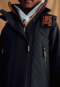 Giacca nera con zip arancioni e un logo ricamato "SUPERDRY" sulla spalla. Presenta diverse tasche con zip e una texture a costine.