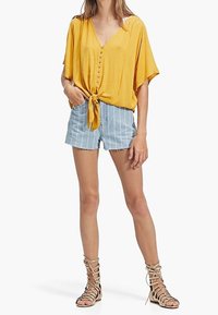 Top giallo testurizzato con nodo frontale, dettagli a bottoni, abbinato a pantaloni corti in denim a strisce blu chiaro e sandali beige con cinturini.