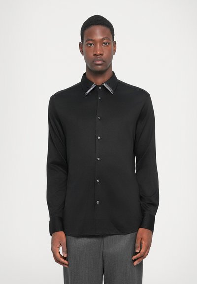 Camisa negra de manga larga con un suave brillo, con cuello puntiagudo con ribete en contraste y cierre de botones, hecha de material suave.