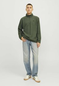 Pullover in pile verde con colletto a mezza zip. Presenta un piccolo logo nero sul petto. Abbinato a jeans azzurri chiari e sneakers bianche.