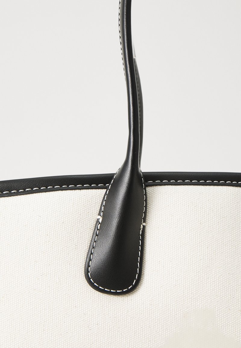 Sac fourre-tout en toile avec un corps blanc, des accents en cuir noir et des coutures. Dispose d'une seule grande anse pour le transport.