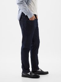 Pantalones vaqueros de mezclilla azul oscuro con corte recto, con costuras naranjas y bolsillos laterales, combinados con una camiseta blanca y una camisa a rayas grises.