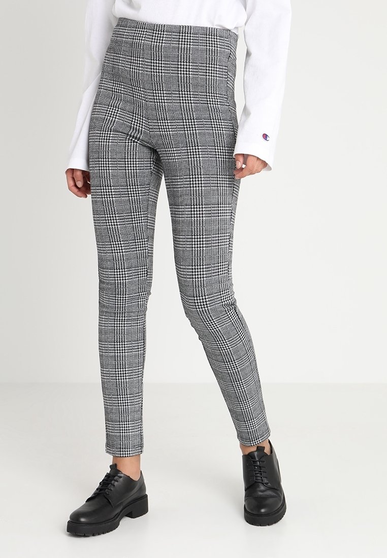 Osoba v šedých a černých kostkovaných slim kalhotách s černými šněrovacími botami a bílém tričku s dlouhým rukávem, na kterém je logo na rukávu.