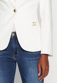 Blazer blanco a medida con detalle de hardware dorado en el bolsillo. Llevado sobre jeans de denim azul, mostrando una tela elegante y un diseño ajustado.