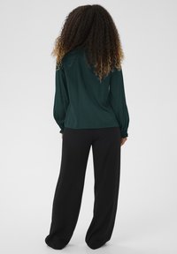 Donna con capelli ricci ombré, che indossa una blusa a maniche lunghe verde scuro e pantaloni neri larghi, in piedi con la schiena rivolta in avanti su uno sfondo semplice.