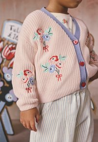 Bērns valkā rozā adītu jaku ar ziedu izšuvumiem un sirds formas pogām, kopā ar gaiši zili baltām svītrainām biksēm.