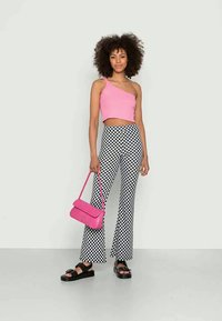 Mulher com top cropped rosa de um ombro só, calças xadrez preto e branco, sandálias plataforma pretas, segurando uma bolsa rosa contra um fundo simples.