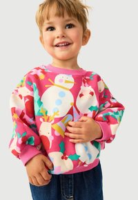 Rosa Fleece-Sweatshirt mit bunten Mustern von Schneemännern und Rentieren. Runder Halsausschnitt, gerippte Bündchen und lockere Passform. Über dunklen Jeanshosen getragen.