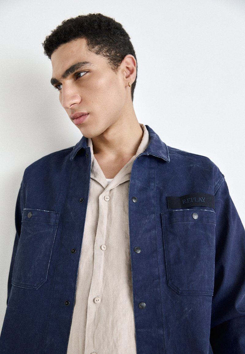 Jeansowa kurtka w ciemnoniebieskim kolorze z zatrzaskami, kieszenią na piersi i czarną metką. Pod spodem widać jasnobeżową koszulę zapinaną na guziki.