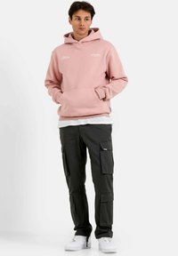 Sudadera con capucha de color rosa claro, con bolsillo tipo canguro, llevada sobre una camisa blanca. Combinada con pantalones cargo negros que tienen bolsillos laterales y zapatillas deportivas blancas.
