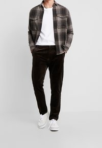 Man wearing white sneakers, dark brown corduroy pants, white t-shirt, and open brown and black plaid shirt with hands in pockets.