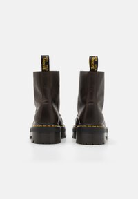 Botas de piel marrón hasta el tobillo con una suela negra gruesa, costuras amarillas y una lengüeta amarilla corta en la parte posterior. Textura del exterior plana y resistente.