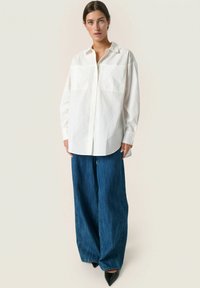 Hvid button-up skjorte med to brystlommer, parret med brede blå denim bukser og sorte spidse hæle. Enkel og struktureret design.