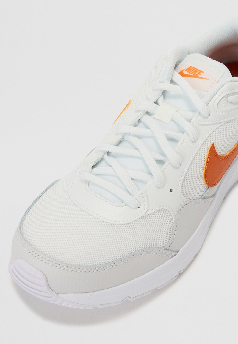 Zapatilla blanca con acentos naranjas, textura de malla y cuero, con punta redonda y sistema de cordones, que presenta un logo en la lengua y en el costado.