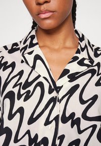 Camicia a righe in bianco e nero con colletto, caratterizzata da linee ondulate audaci su uno sfondo chiaro e piccoli bottoni frontali.