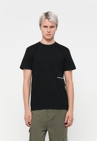 Zwarte katoenen T-shirt met korte mouwen, ronde hals, met witte tekst "weekend deelnemer" aan de linksonderkant. Effen en ontspannen pasvorm.