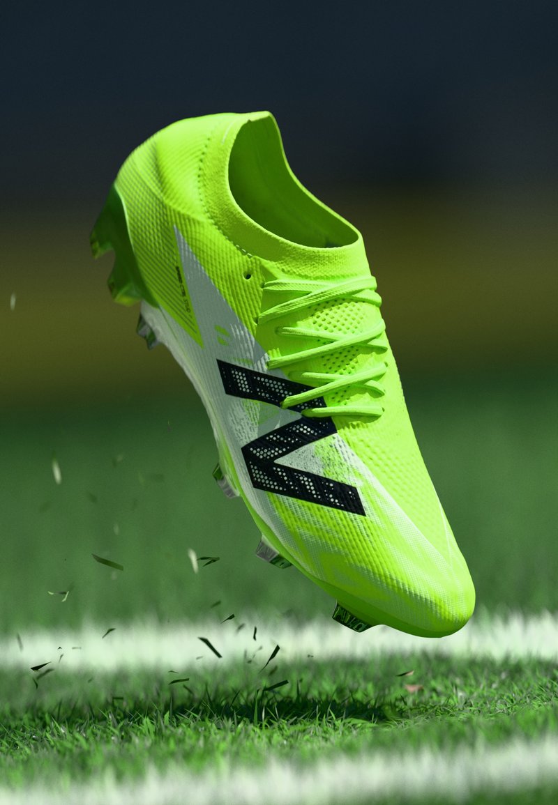 Crampon de football vert citron avec une surface texturée, un motif noir accentué, et six crampons, suspendu au-dessus de l'herbe avec des débris volant.