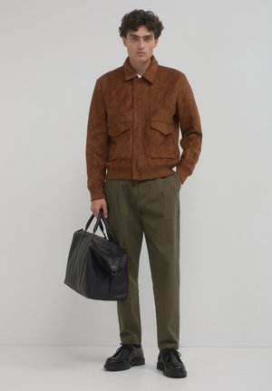Veste bomber en suède marron avec deux poches avant, pantalon vert olive et sac noir. Le mannequin se tient avec les cheveux coiffés en douces boucles.