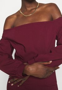 Burgunderfarbenes Off-Shoulder-Oberteil mit langen, gepufften Ärmeln und tailliertem Oberteil. Eine goldene, geschwungene Halskette akzentuiert den Ausschnitt. Glatte Stofftextur.