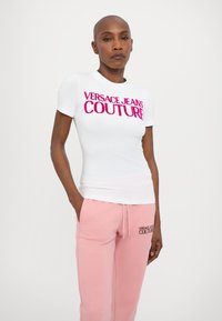 Valge lühikeste varrukatega T-särgi, millel on roosa "VERSACE JEANS COUTURE" logo. Kombineeritud helesiniste dressipükstega, millel on sarnane logo tikand.