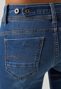 Pantalones vaqueros azules de denim con un bolsillo trasero que presenta costuras naranjas, botones metálicos en la cintura y un ajuste tapered clásico.