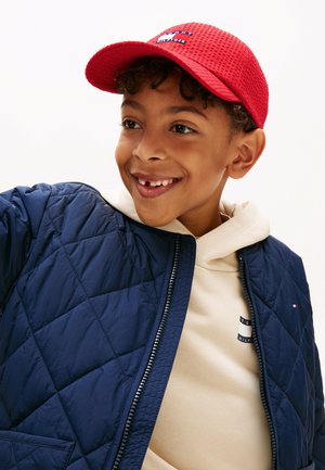 Naeratav poiss, kellel on peas punane nokamüts, beež kapuutsiga dressipluus ja tumesinine tekkidest jope, vaatab küljele tühja heleda tausta ees.