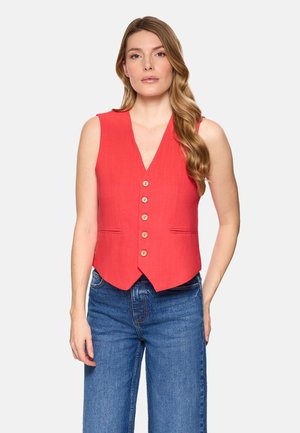 Mujer con cabello largo y ondulado, vestida con un chaleco rojo sin mangas con botones y jeans azules, de pie frente a un fondo claro y liso.