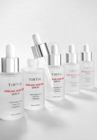 Cinque bottiglie in vetro smerigliato di siero all'acido azelaico 12% di TirTir con tappi argentati, un contagocce sollevato da cui sta cadendo una goccia.