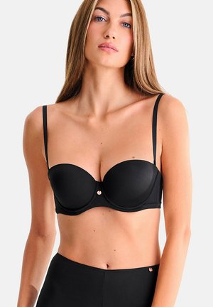 Femme aux longs cheveux portant un soutien-gorge noir sans bretelles et un bas noir taille haute, vue de face sur un fond blanc uni.