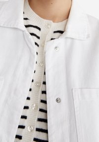 Chaqueta de mezclilla blanca con botones de presión plateados, superpuesta sobre un suéter crema con rayas horizontales negras y botones redondos.