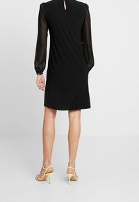 Frau in einem knielangen schwarzen Kleid mit transparenten langen Ärmeln und gelben High-Heels, die vor einem schlichten hellen Hintergrund steht.