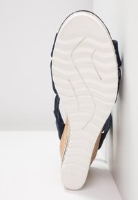 Semelle blanche d'une sandale compensée avec talon en liège et brides bleu marine, vue du dessous sur une surface et un fond blanc.
