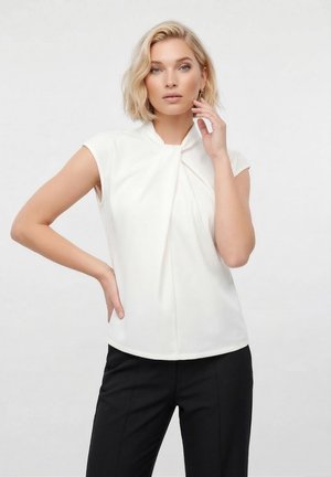 Ξανθιά γυναίκα με κοντά μαλλιά, φοράει λευκό αμάνικο τοπ και μαύρο παντελόνι, ποζάροντας με το ένα χέρι στη μέση και το άλλο κοντά στο πρόσωπο.