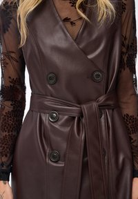 Kobieta w ciemnobrązowej skórzanej sukience z paskiem noszonej na przezroczystej czarnej koszulce z długim rękawem w kwiatowy wzór, eksponująca szczegóły torsu i talii.