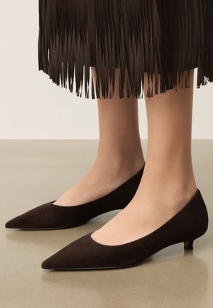 Füße in dunkelbraunen spitzen Wildleder-Kitten-Heels mit einem dunkelbraunen Fransenrock, der oberhalb der Knöchel sichtbar ist.