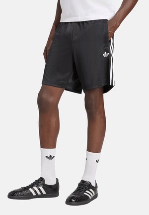 Persona que lleva pantalones cortos negros de Adidas con rayas blancas, calcetines blancos de Adidas y zapatillas negras de Adidas, de pie con las manos en los bolsillos.