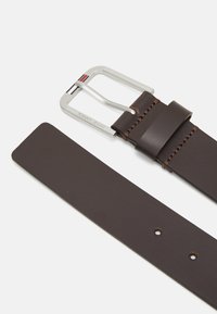 Cinturón de cuero marrón con hebilla de metal plateado, estampada con "TOMMY JEANS", y detalle de costura contrastante. Textura suave y forma rectangular.
