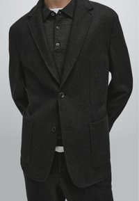 Dunkelgrauer Woll-Mix-Blazer mit strukturierten Schultern, Reverskragen, zwei aufgesetzten Taschen vorne und Knopfverschluss. Getragen über einem passenden Poloshirt.