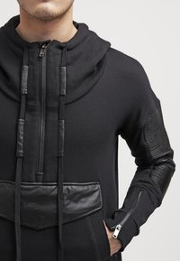 Sudadera negra con capucha, que presenta un cierre con cremallera en el frente, acentos de piel texturizada y mangas acanaladas. Los gruesos cordones y el cuello alto añaden un toque de detalle.