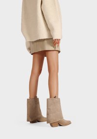 Stivaletti in suede beige con tacco a blocco, caratterizzati da un design strutturato e cuciture sottili. Indossati con una gonna chiara e un maglione over size.