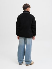 Black fleece jacket with a high collar, worn over a black shirt. Paired with loose light blue jeans and beige footwear.