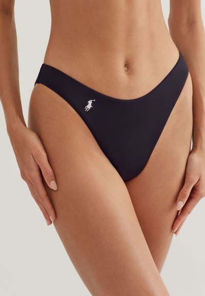 Γυναίκα που φοράει μαύρο κάτω μέρος μπικίνι με μικρό λευκό λογότυπο παίκτη πόλο στο αριστερό ισχίο, τα χέρια ακουμπούν στα πλάγια, ουδέτερο φόντο.