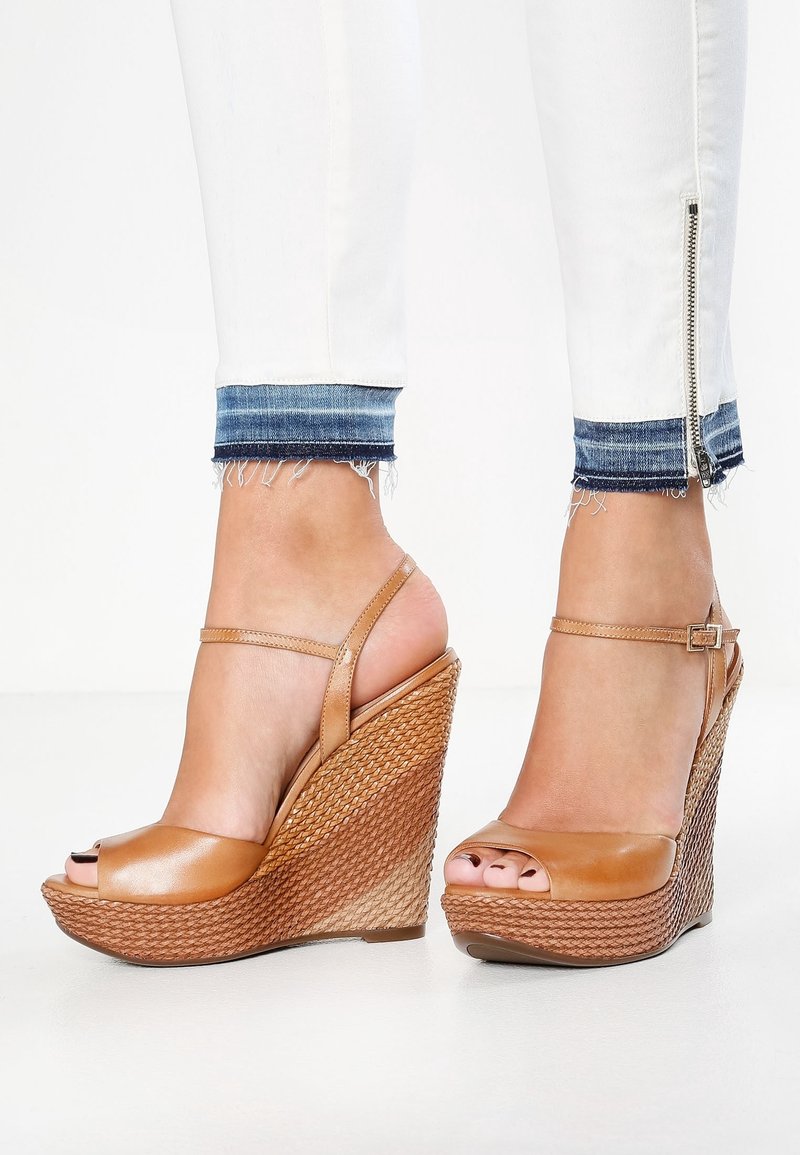 Pieds portant des sandales compensées en cuir marron clair avec des brides à la cheville, associées à un jean blanc avec des revers en denim et des fermetures éclair latérales.