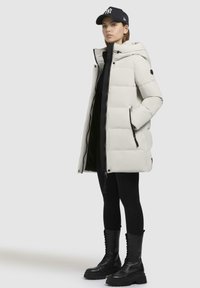 Giacca a vento in bianco sporco, con cappuccio, chiusure a zip e tasche laterali. Indossata con leggings neri e stivali neri con lacci spessi.