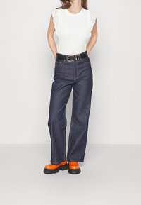 White top with eyelet trim sleeves, dark blue high-waisted jeans, and chunky orange platform shoes. Black belt with shiny hardware accents.