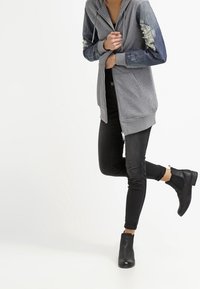 Felpa grigia con zip e maniche in denim, dettagli strappati sulle maniche, indossata con jeans skinny neri e stivaletti neri.