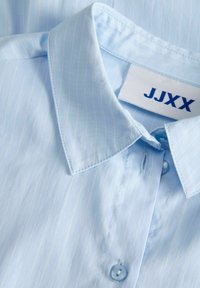 Col de chemise bleu clair à fines rayures avec boutons transparents et une étiquette blanche affichant "JJXX" en texte bleu foncé.