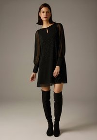 Schwarzes Kleid mit langen, durchsichtigen Ärmeln, Ausschnitt mit Schlitz und mit kleinen Punkten verziert; kombiniert mit schwarzen Overknee-Stiefeln.